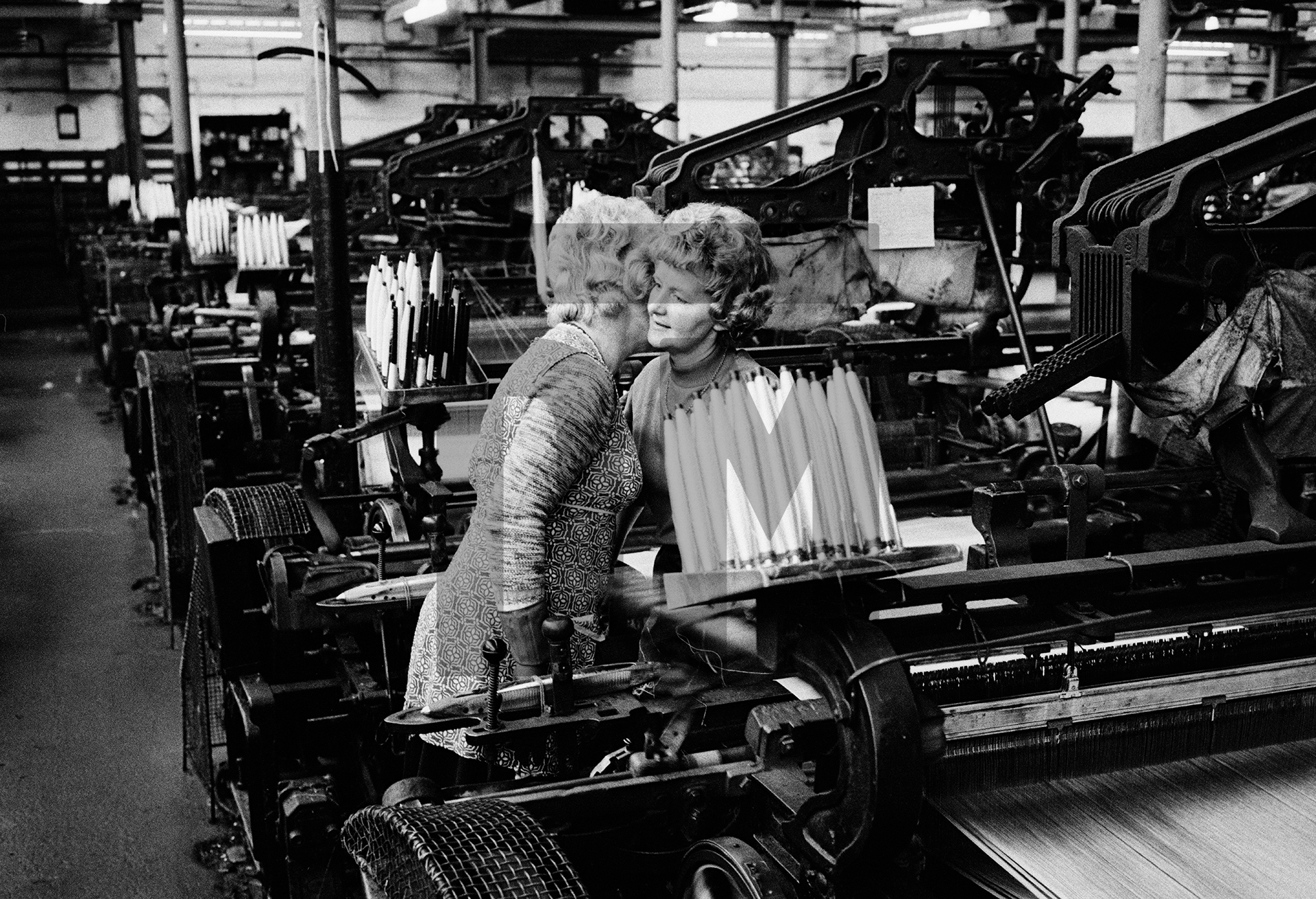 Two weavers in conversation, their faces close together so they can chat privately above the 'clatter' (deafening noise) of the looms without others nearby, skilled lip-readers all, being able to follow their conversation, Spring Bank Mill, Fence. Autumn 1975 by Daniel Meadows